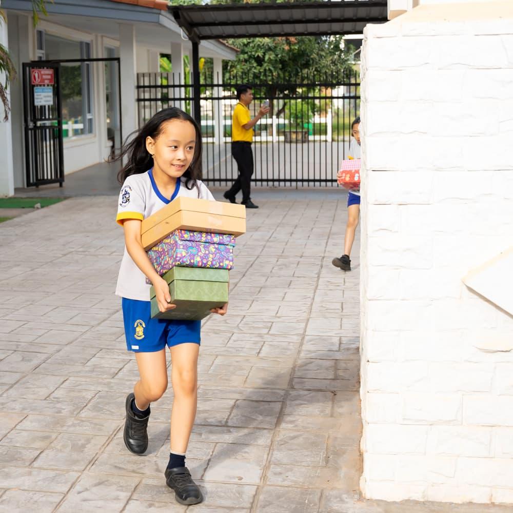 A student carrying colorful gift boxes across the school courtyard during the Holiday Box Project.