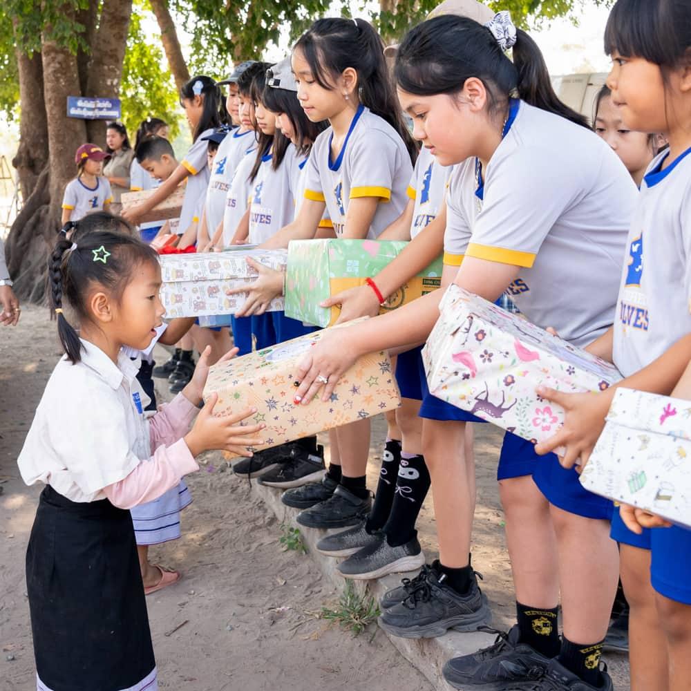Students from the school lined up, each offering a wrapped gift box to younger children in a heartfelt exchange.