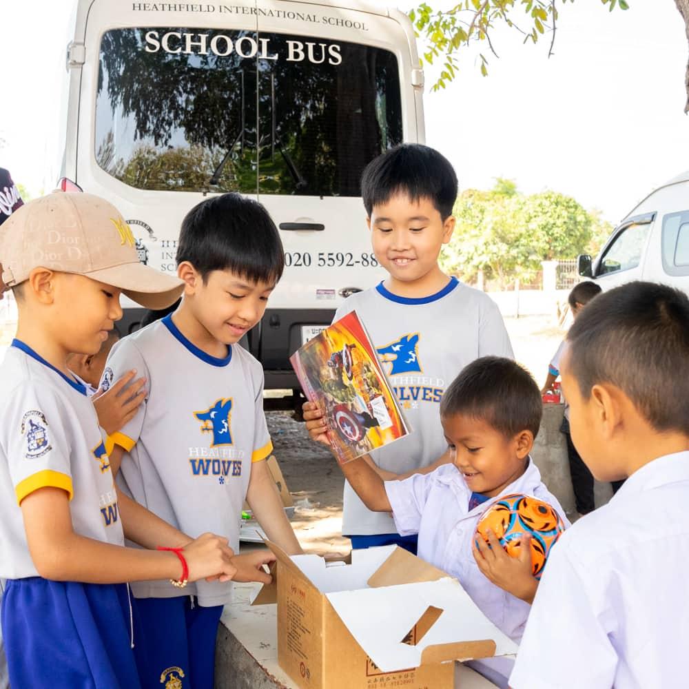 A group of students gathered near the school bus, opening a box to reveal gifts while sharing smiles and excitement.