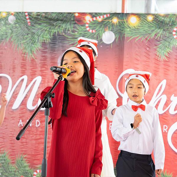 A student in a red dress stands at the microphone as the lead singer, accompanied by a choir of students wearing coordinating Christmas attire.