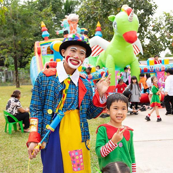 A cheerful clown interacts with a child dressed as an elf, with colorful inflatables and activities in the background adding to the festive atmosphere.