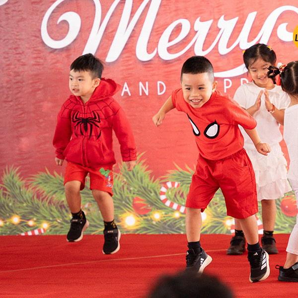 Young students dressed in red, including Spider-Man-themed outfits, energetically perform a lively routine in front of a holiday backdrop.
