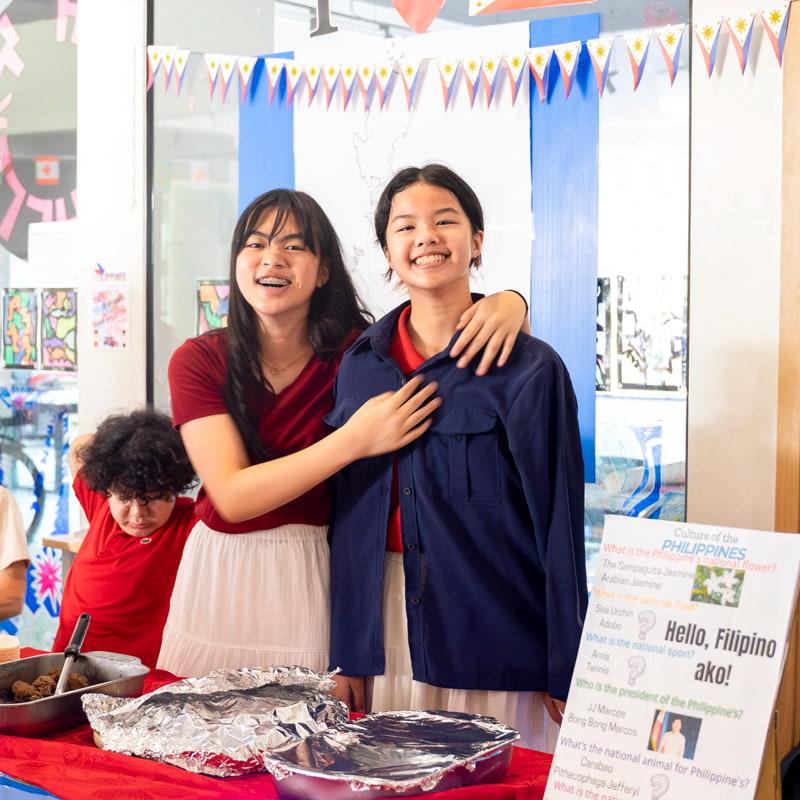 Two girls standing by a Filipino cultural booth, smiling and serving food.