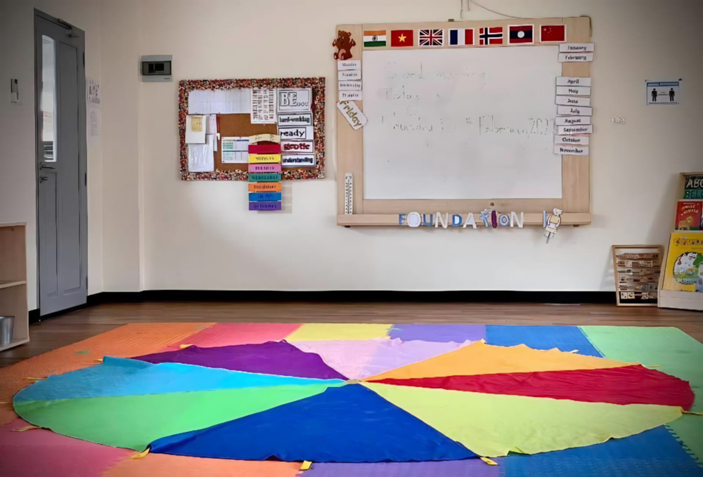 Colorful early years classroom in an international school, featuring a bright circular floor mat, educational displays, and flags from various countries above the whiteboard.