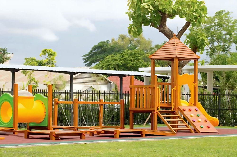 An Early Years Foundation Stage (EYFS) playground with a lush green lawn, colorful play equipment, and shaded areas under a canopy of flags, designed for young children.
