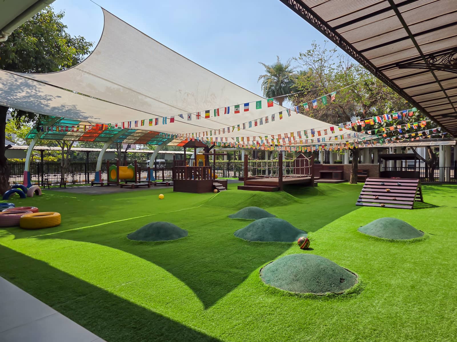 An Early Years Foundation Stage (EYFS) playground with a lush green lawn, colorful play equipment, and shaded areas under a canopy of flags, designed for young children.
