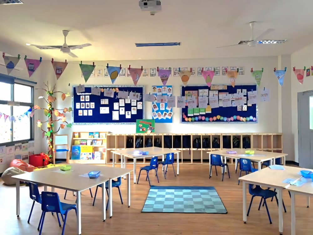 A bright and colorful primary classroom with decorated walls, flags hanging from the ceiling, and neatly arranged desks and chairs.